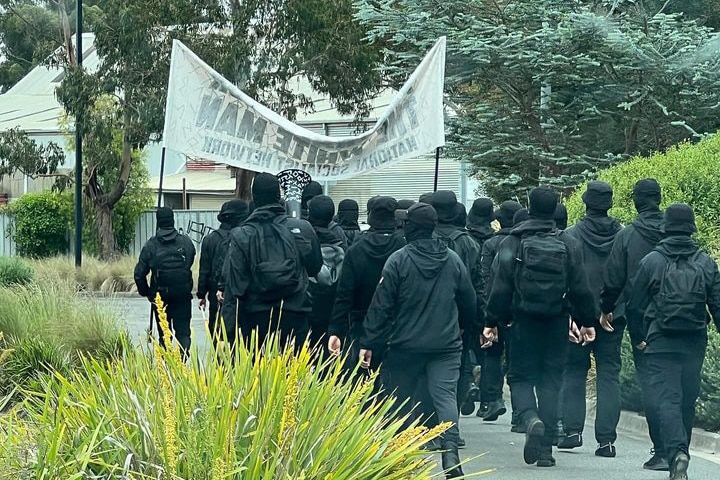 A group of men in black wearing balaclavas walk behind a banner.