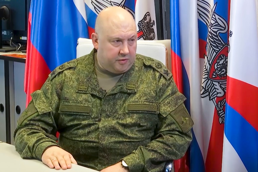 一位身穿军装的重量级秃头男子坐在俄罗斯国旗变体前的椅子上。