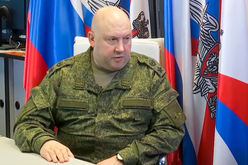 A heavy, bald man in military fatigues sits in a chair in front of a variation of the Russian flag.