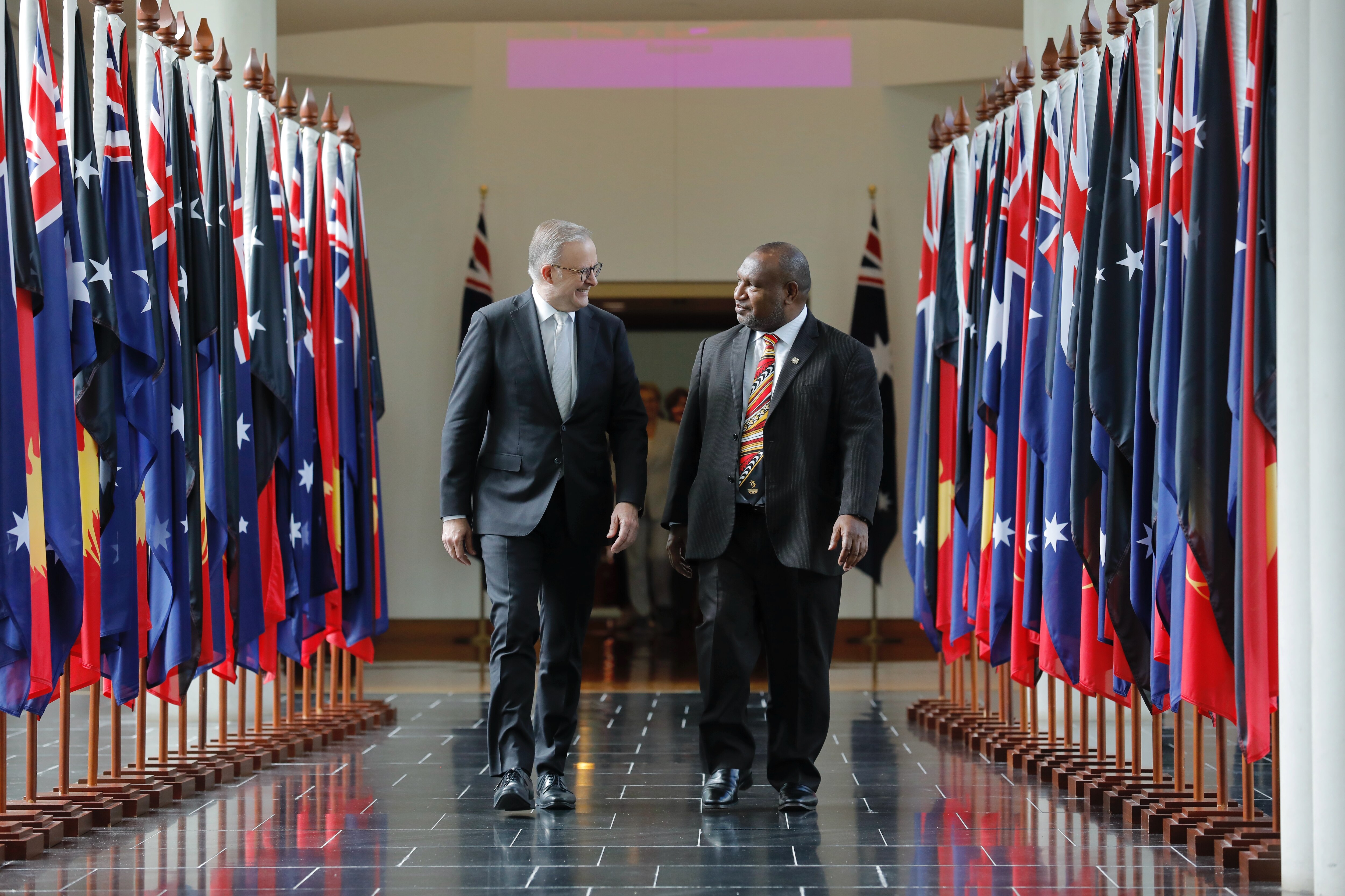 Two men wearing suits walking in a hallway lined with flags