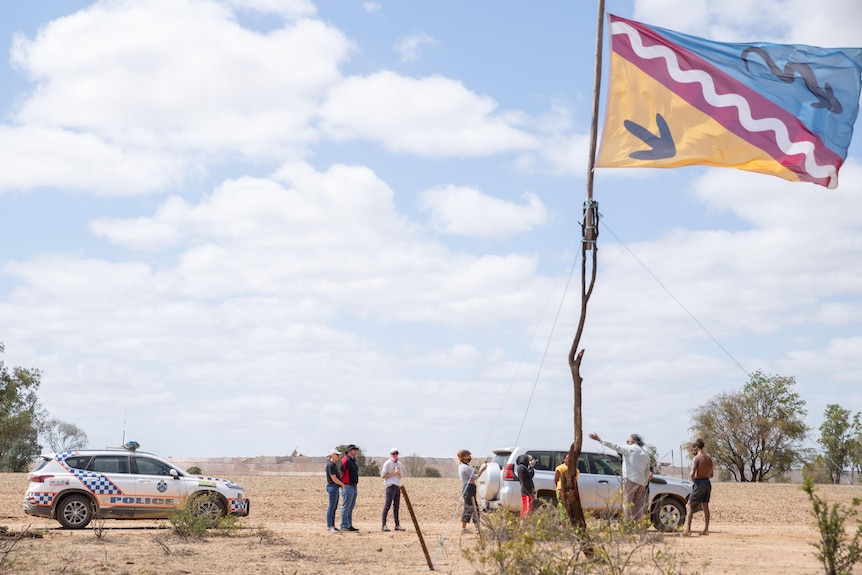 people stand around a police car talking. there is a symbolic flag flying in the foreground