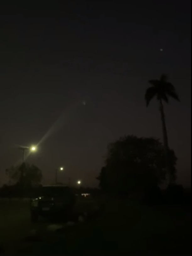 A small bright white dot is seen in against the night sky, a palm tree silhouetted in front