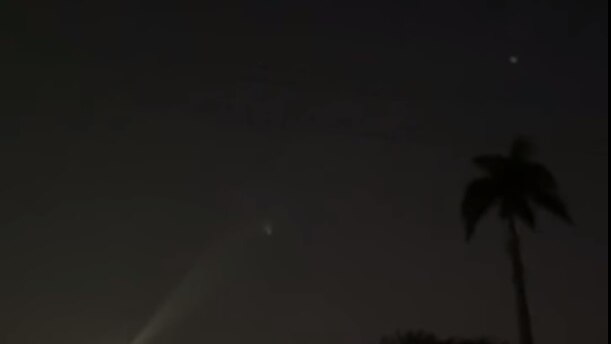 A small bright white dot is seen in against the night sky, a palm tree silhouetted in front