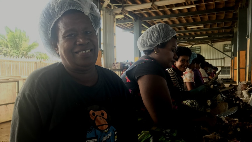 A woman smiles at the camera with a knife and casava in her hands and other workers sitting behind her in a factory.