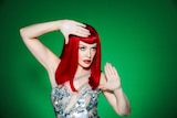 A red-haired Kylie Minogue poses with her hands, wearing a a glittery frock against a dark green background