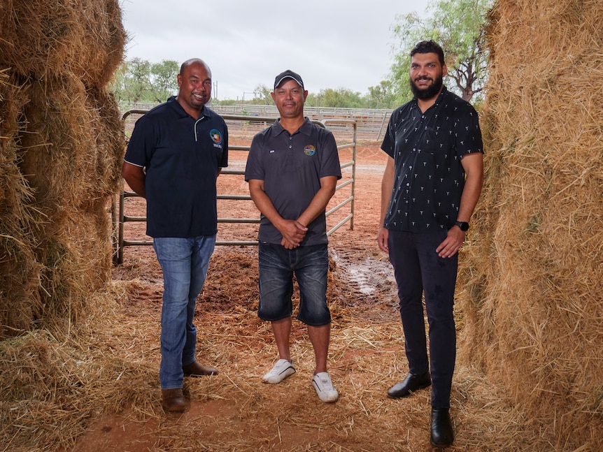 Three indigenous men stand next to hay bales
