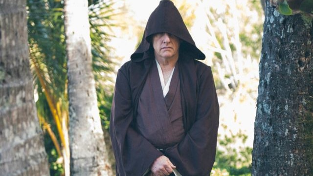 Man stares at the camera with a stern expression while dressed in Jedi knight robes including a black hooded cloak.