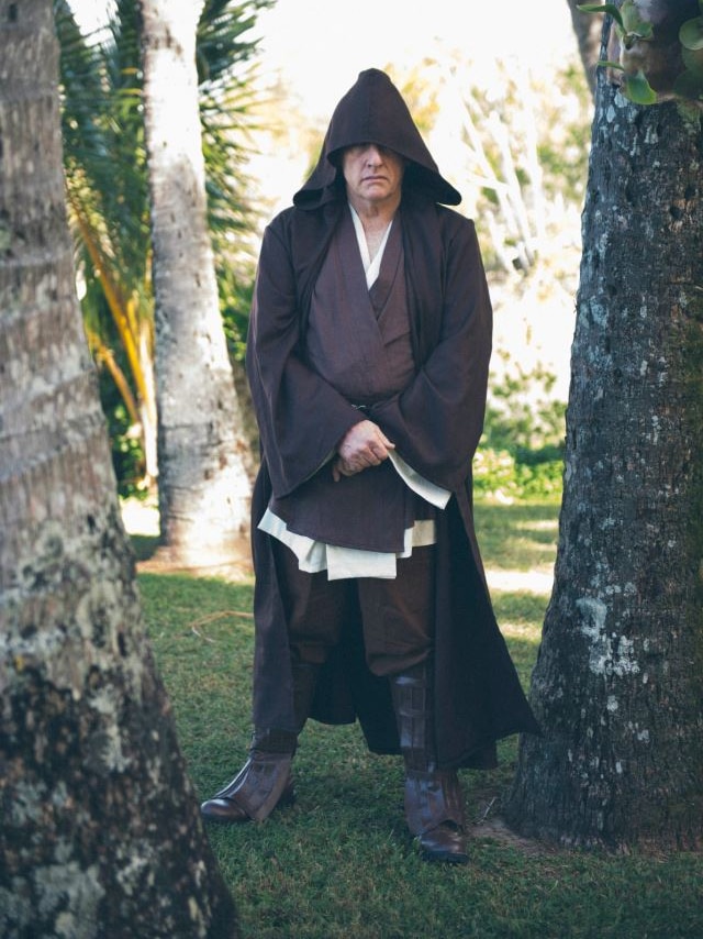 Man stares at the camera with a stern expression while dressed in Jedi knight robes including a black hooded cloak.