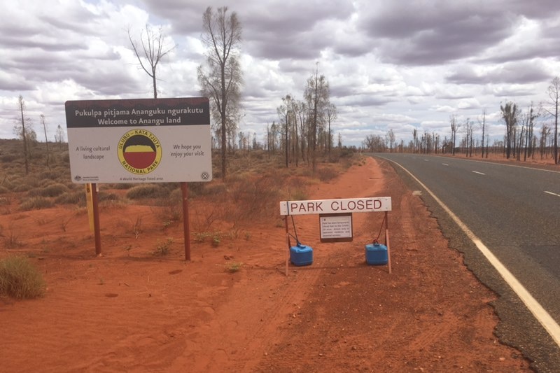 Two signs in red dirt next to bitumen road. Grey clouds in sky