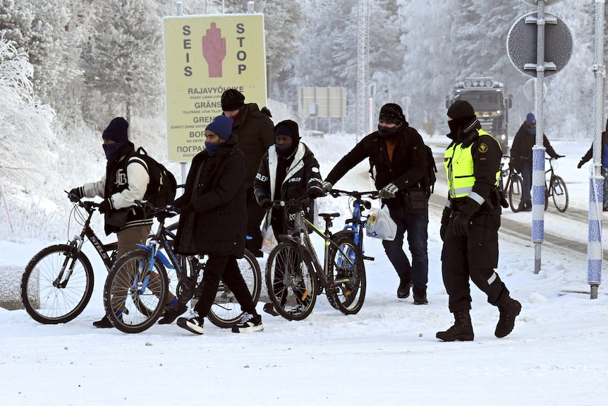 Seven men walk their bicycles in the snow, two border guards with high-vis vests escort them