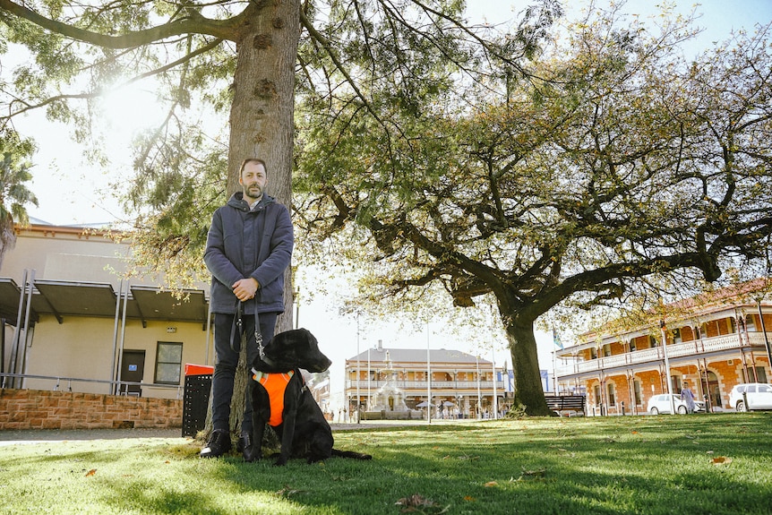 A man leans against a park tree holding the leash to a seated black dog wearing an orange vest.