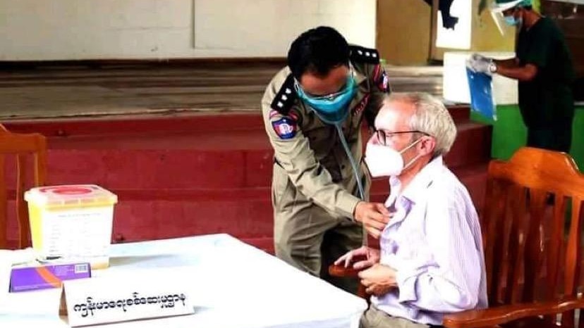 A man wearing a pink shirt and mask sits in a chair while a doctor checks his chest.