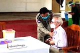 A man wearing a pink shirt and mask sits in a chair while a doctor checks his chest.