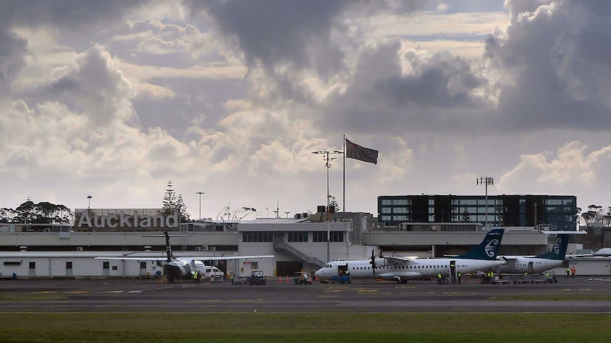 A wide view of Auckland Airport from the outside. A New Zealand flag flies on top of the building and planes are on the tarmac.