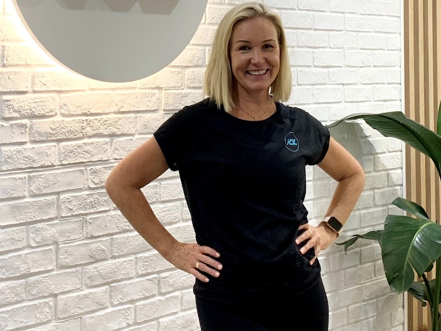 A blonde lady smiles at the camera with her hands on her hips in front of a white brick wall