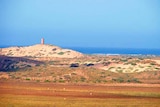 A vista of sand dunes, covered in shrubs, near the sea.