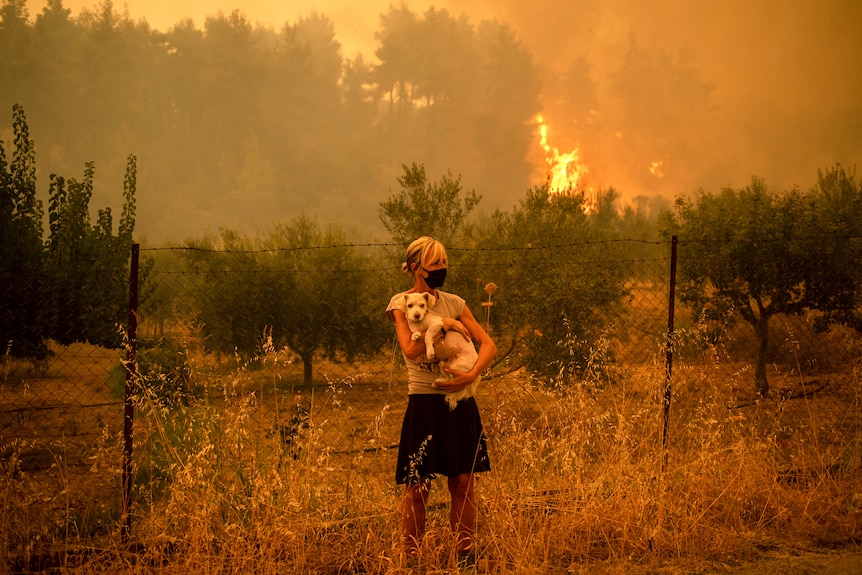 A masked woman holds her dog in her arms as flames leap in the forest behind her