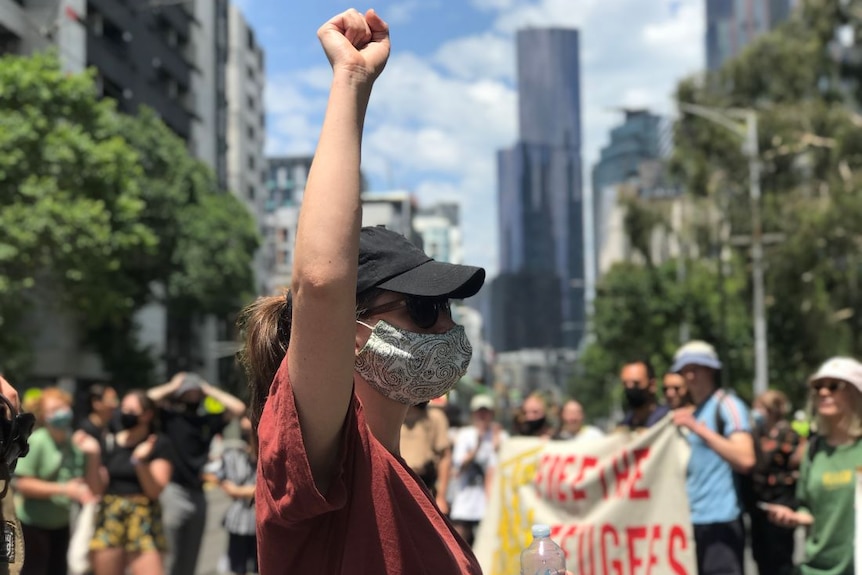 A woman in a red t-shirt, black cap and face mask raises a clenched fist in the air as people gather behind her.