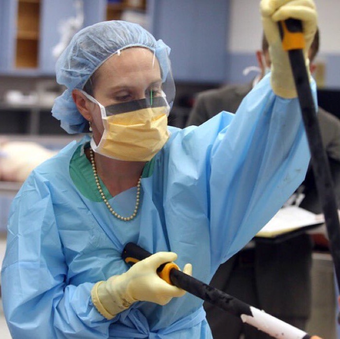 A doctor in scrubs and a face mask prepares to use a large cutting tool in a morgue
