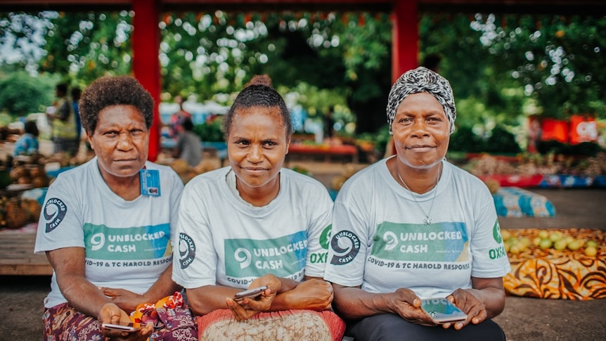 Women hold phones and smile at camera, wearing branded t-Shirts.