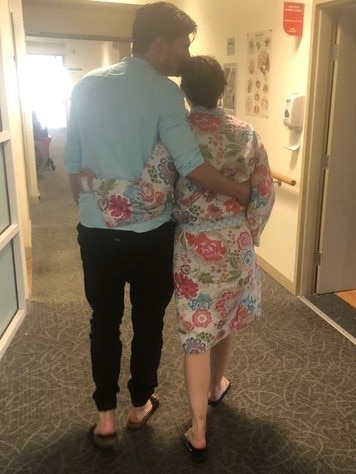 A man and woman walk down a hospital corridor with the arms around each others backs. 