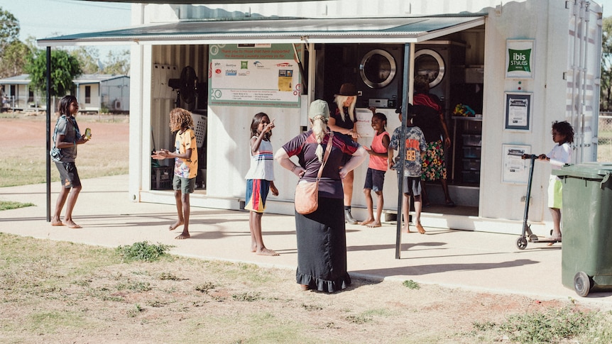 A shipping container housing washing machines has people out the front waiting for their washing to be cleaned. 