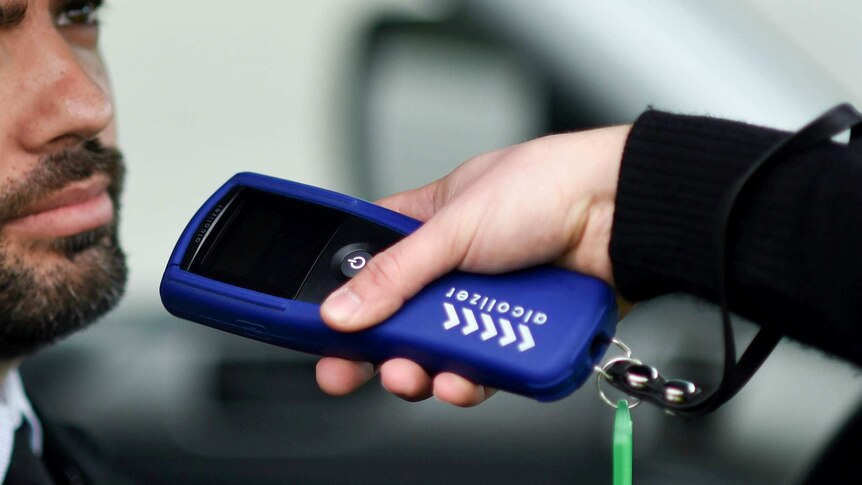 A hand holding a breathalyser is held up to a man's face.