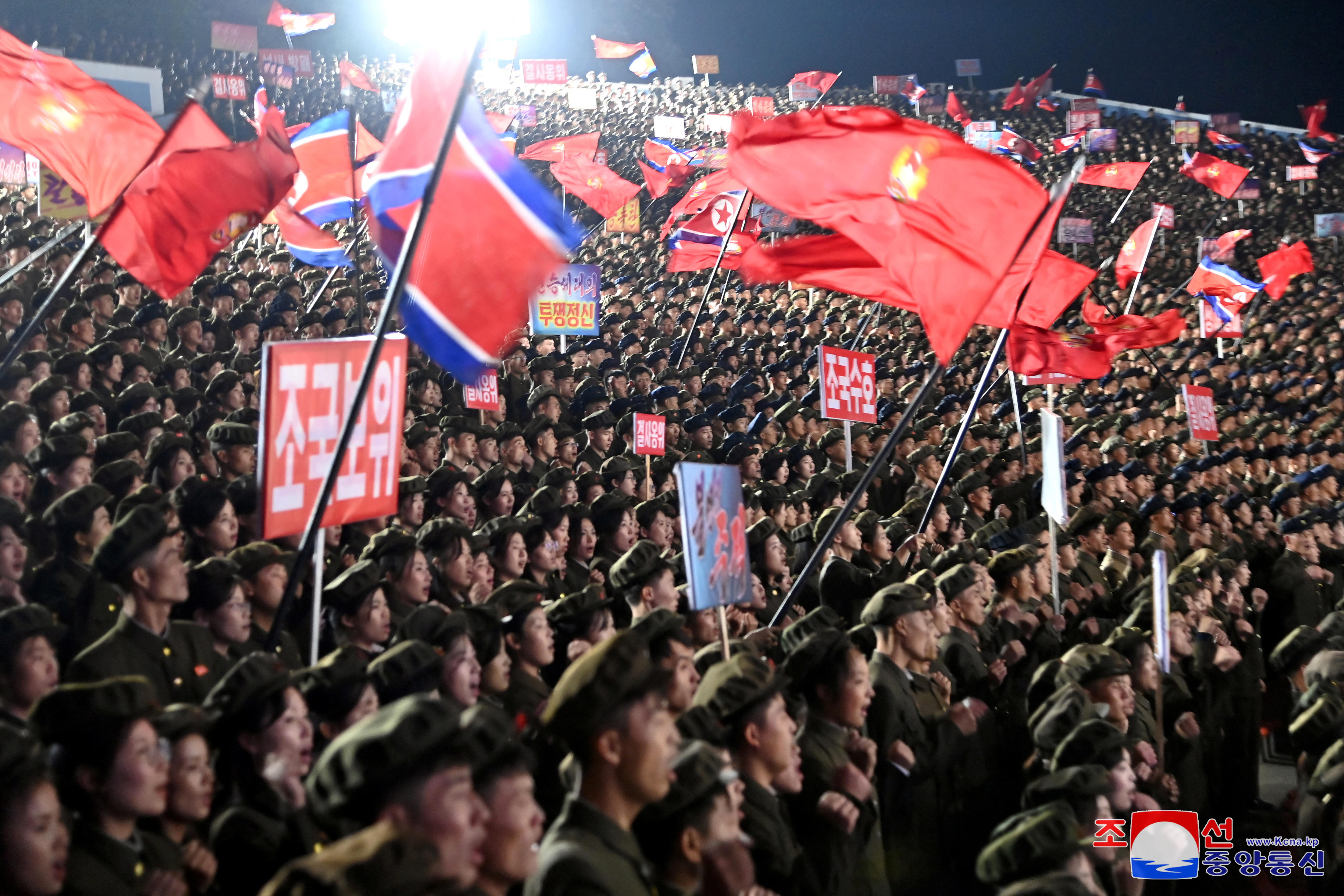 Thousands of people stand in army uniforms waiving North Korean flags in a stadium