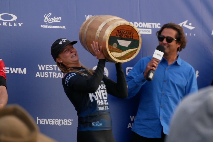 A woman lifting a small wine barrel, about the size of their body, with a man with microphone watching on a stage