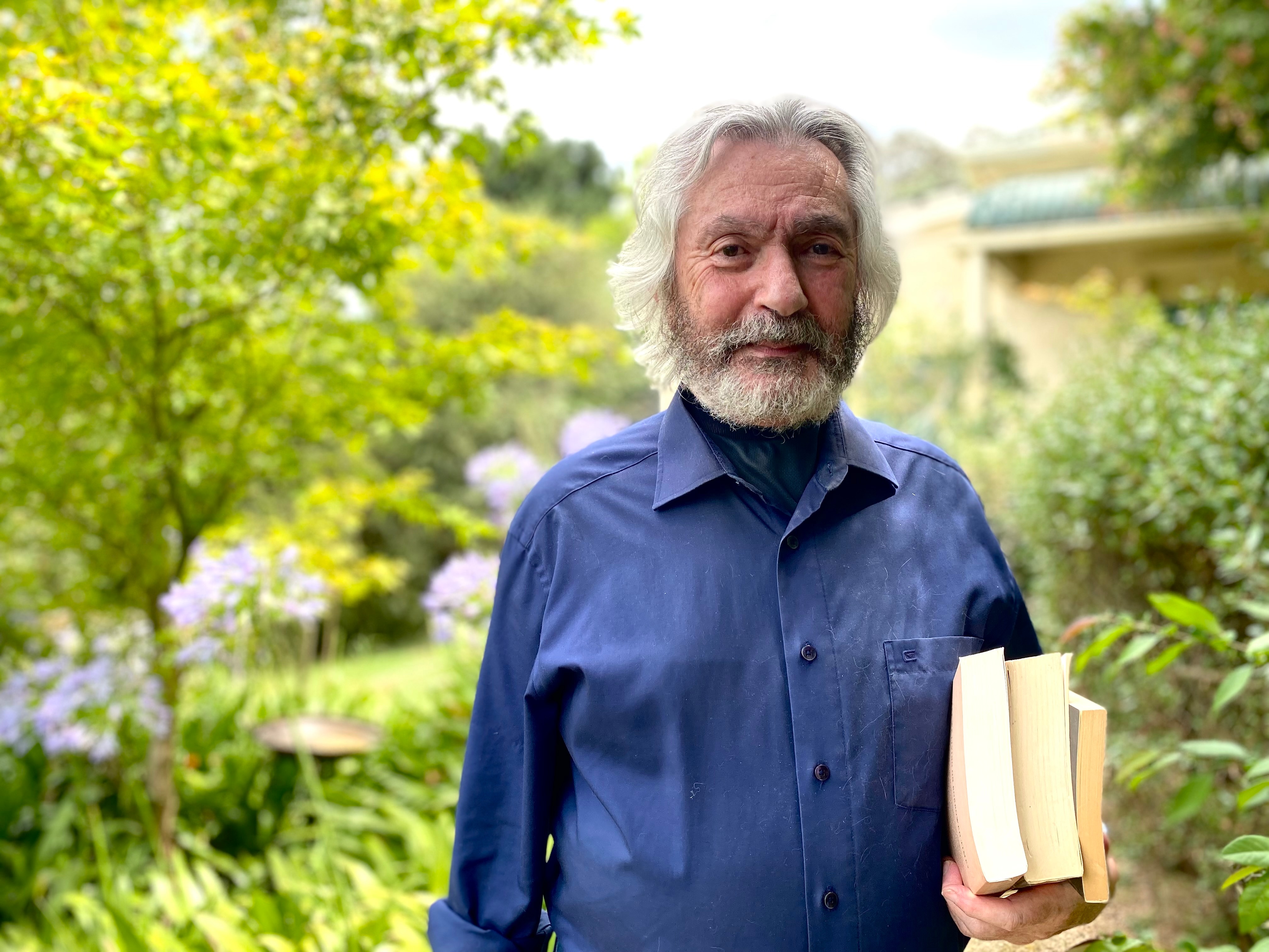 A man with white hair wearing a blue shirt holds three books in his arm