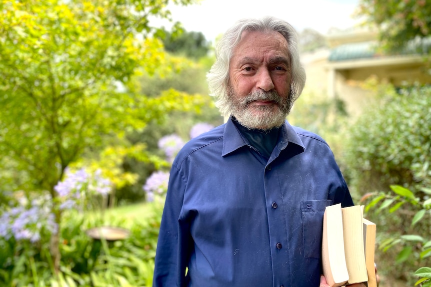 A man with white hair wearing a blue shirt holds three books in his arm