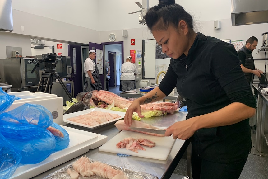 Image of an Asian woman in black work clothes cutting fish in a busy commercial kitchen where filming is taking place.