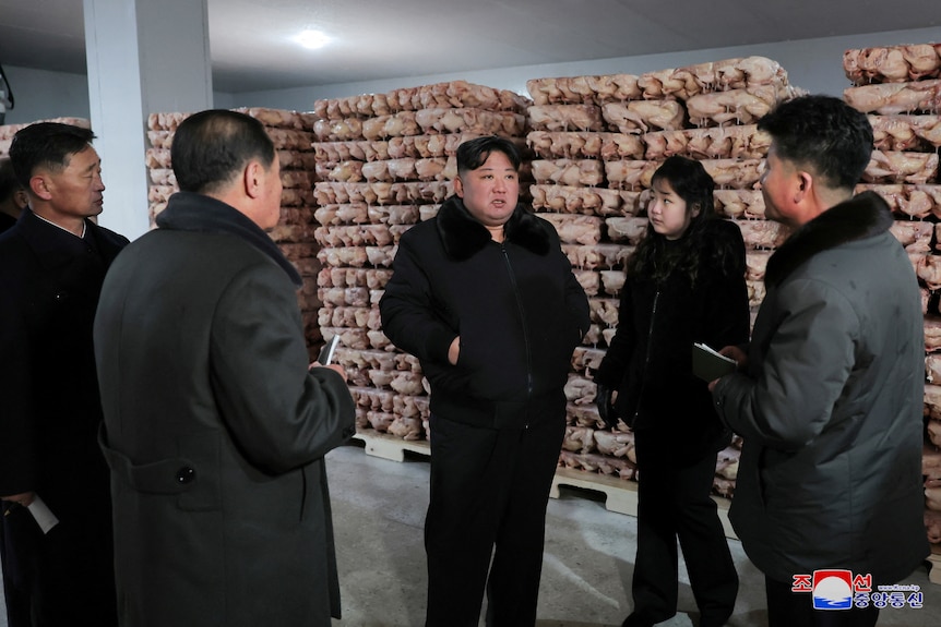A middle-aged Korean man and a young girl, both dressed in black, stand in front of stacks of dead chickens.