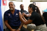 Michael Liddle, a man, gets a COVID vaccine in his left arm, while another man looks on from behind.