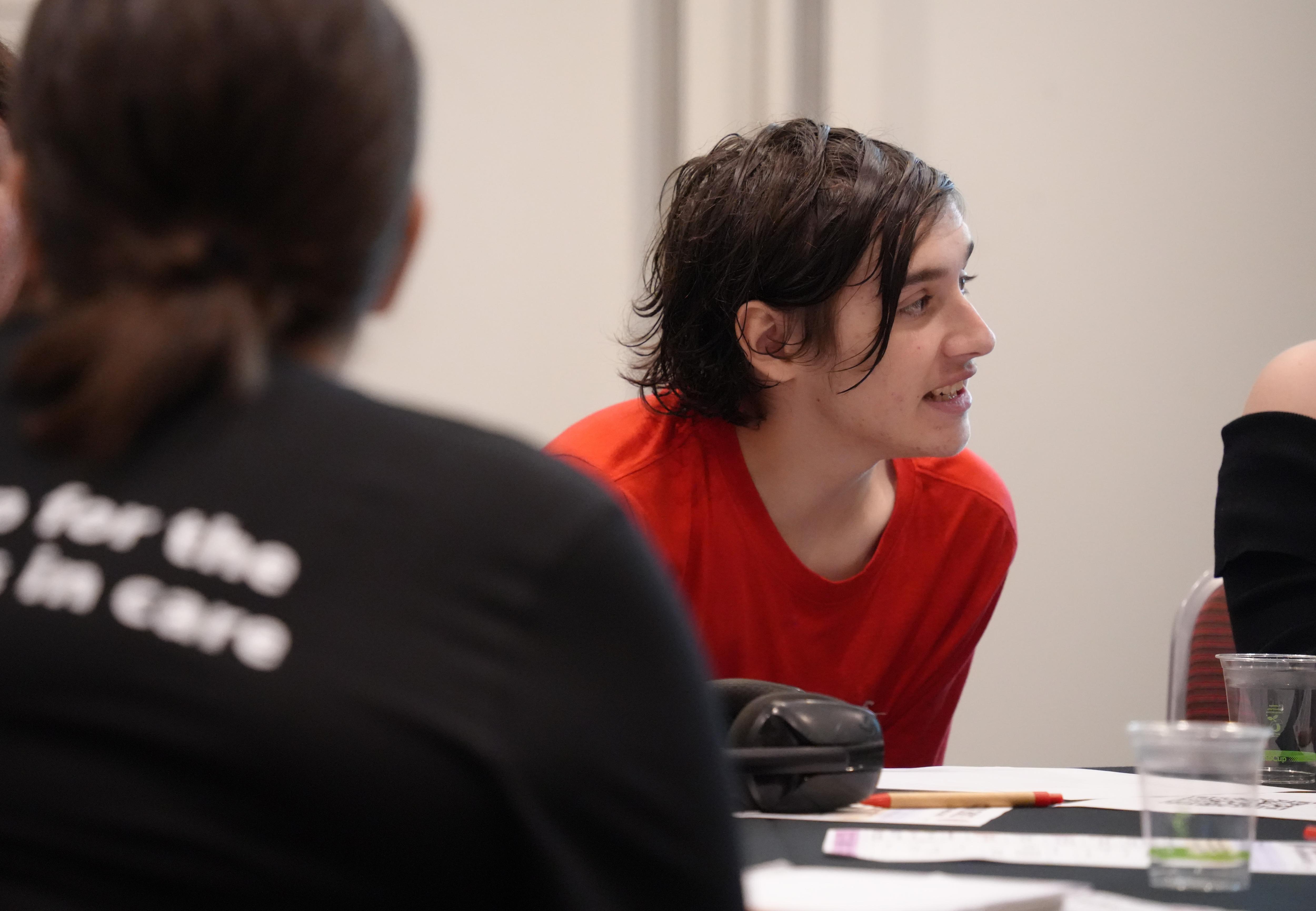 Side-view photo of a  man with brown hair wearing a red t-shirt speaks to people around a table.