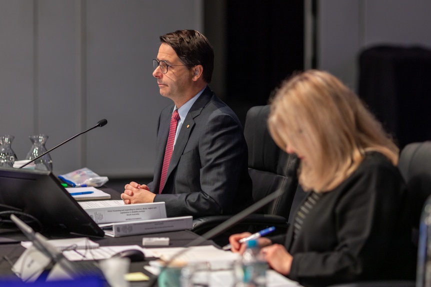 A photo of Alastair McEwin in profile seated at a table with a microphone.