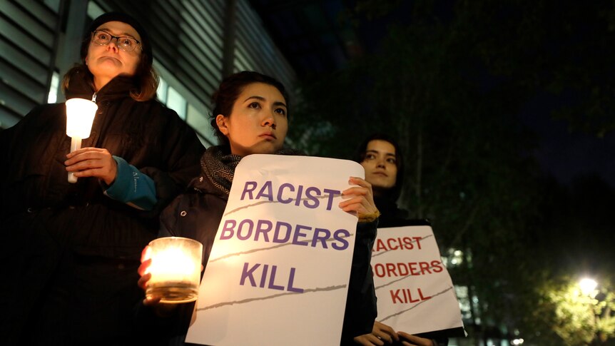 Three women stand together holding lit candles and posters that read "racist borders kill" in all capitals