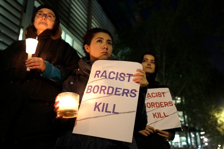 Three women stand together holding lit candles and posters that read "racist borders kill" in all capitals