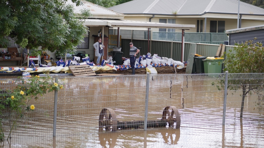 Two people stand in the front yard of a sandbagged home surrounded by floodwater.
