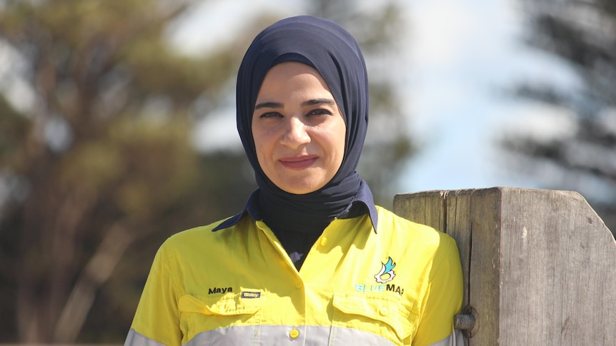 She wears hi vis and leans against a jetty post, with blurred outlines of pine trees behind her