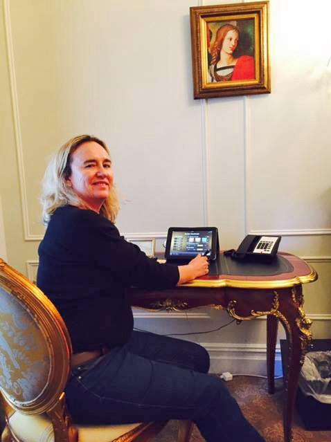 A woman sits at an ornate desk in a hotel writing on a tablet