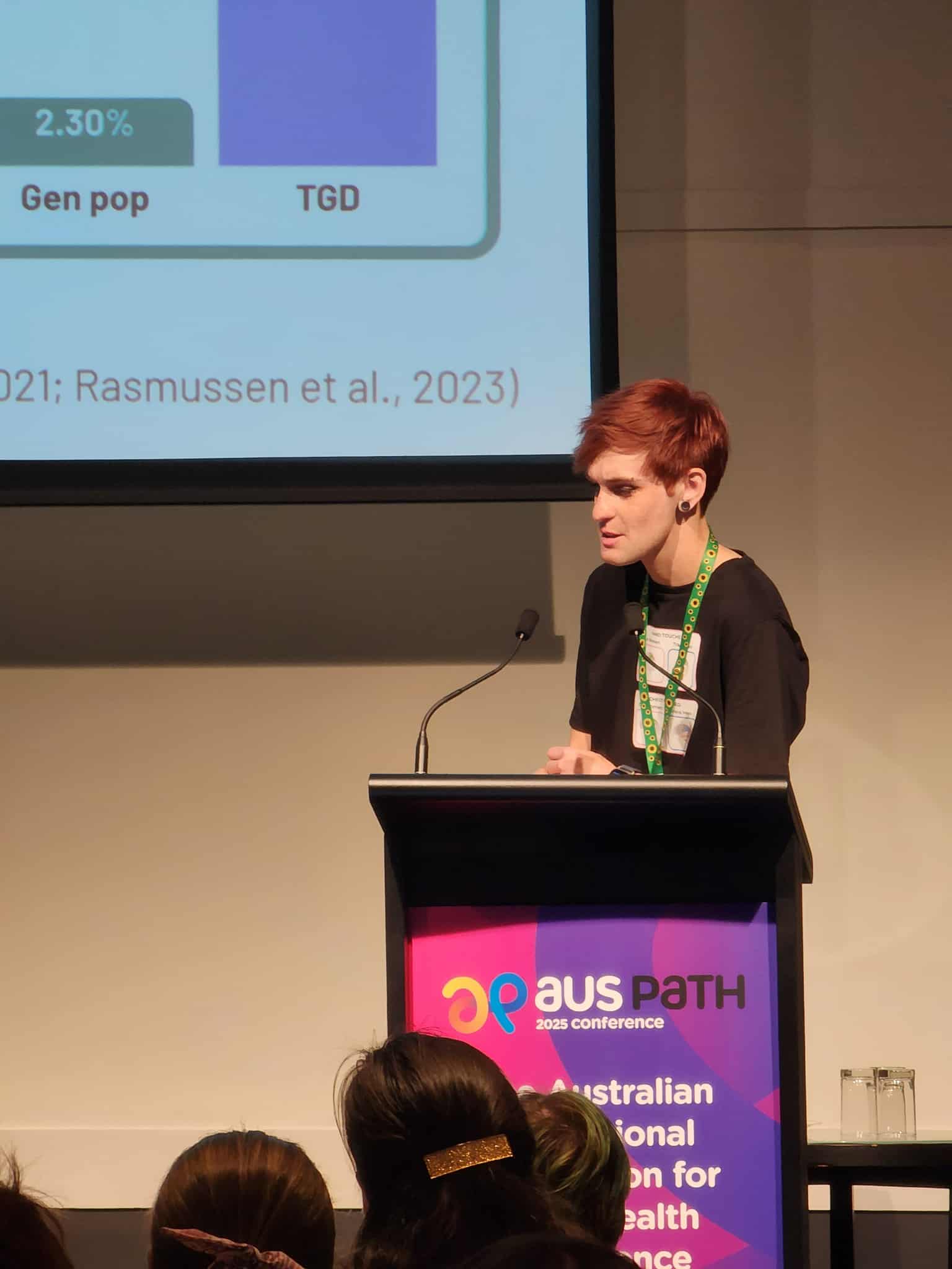 A speaker with short pink hair at a lecturn talks to the audience.
