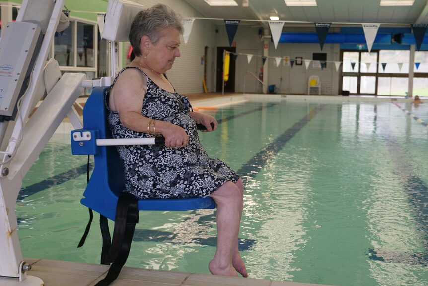 A woman in bathers sitting on a chair that is lowering her into an indoor pool. 