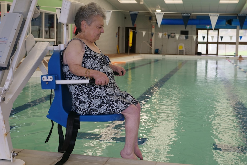 A woman in bathers sitting on a chair that is lowering her into an indoor pool. 