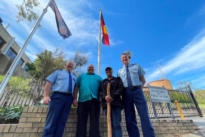 Four men smile in front of the Aboriginal flag.