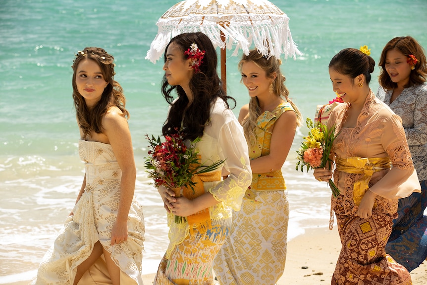Five young women walk on a sunny beach as a bridal party; the bride leads with brown hair and a shoulder-less white dress