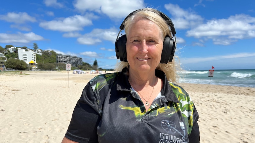A lady with blonde hair and wearing headphones smiles while standing on a Gold Coast beach