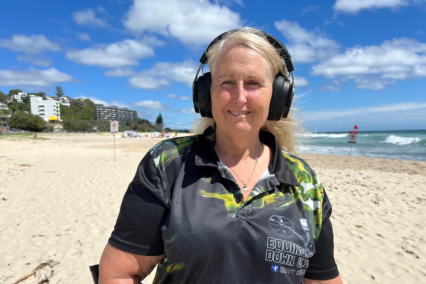 A lady with blonde hair and wearing headphones smiles while standing on a Gold Coast beach