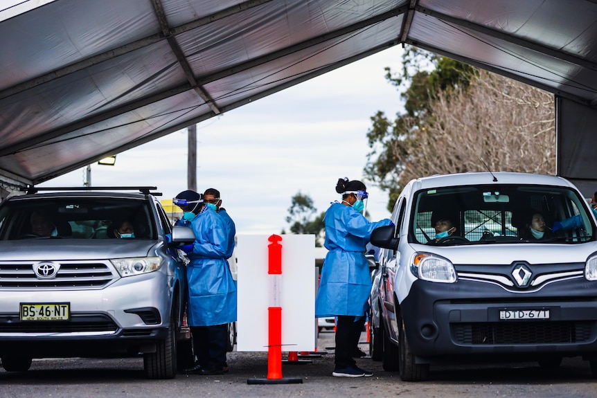 Two staff wearing protective gowns and masks and face shields lean into cars under an awning. 