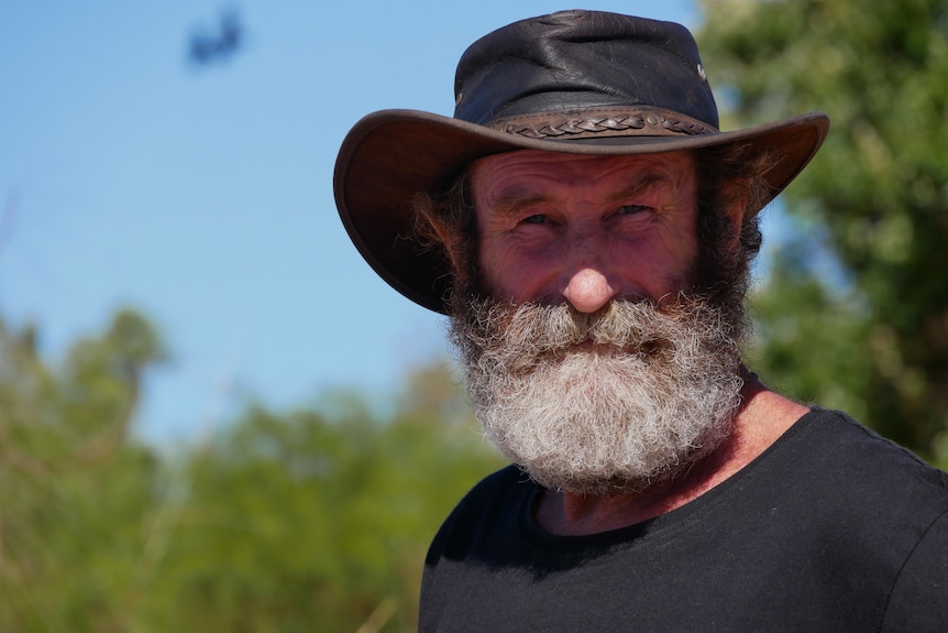 Man with grey beard and hat stares into the camera with sky and bush behind him.